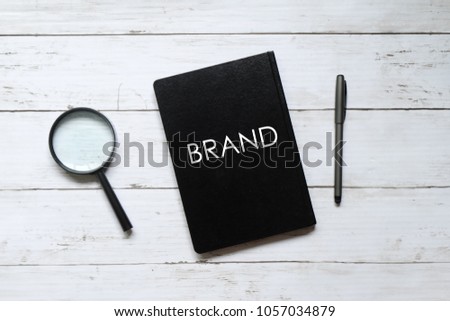Top view of magnifying glass,pen and notebook written with 'BRAND' on white wooden background.