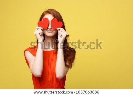 Lifestyle Concept: Attractive woman with beaming smile having two small red hearts in hands, closing eyes with paper heart symbols while standing over yellow background.