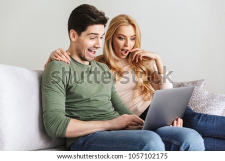 Photo of excited surprised young loving couple sitting on sofa indoors at home using laptop computer.