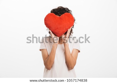 Picture of amusing woman 20s with brown hair covering face with red heart shape pillow isolated over white background