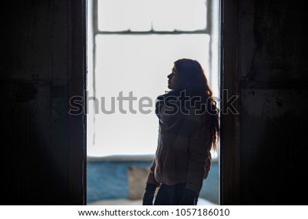 Silhouette of a young cute white girl in warm scarf and gloves is standing opposite window of a ruined house in winter. Destroyed building.