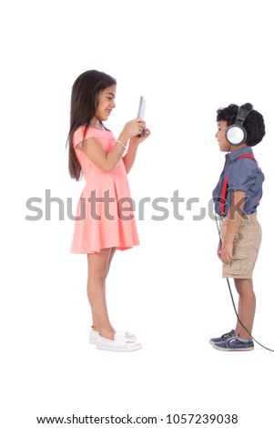 Two kids standing the boy putting the headphone on his head and the girl taking a photo with the mobile, isolated on a white background.