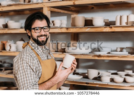 smiling potter holding ceramic cup in workshop with dishware on shelves
