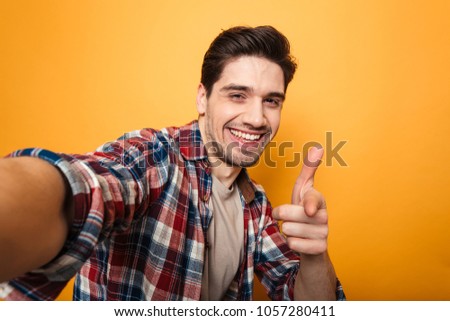Portrait of a cheerful young man taking a selfie and pointing finger isolated over yellow background