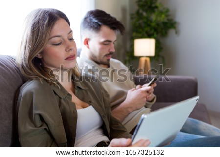 Portrait of beautiful young bored couple using their digital tablet and smartphone at home.