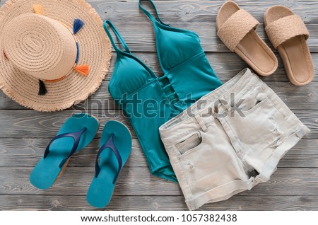 Womens clothing, accessories (denim shorts, straw hat, swimsuit, sandals) on grey wooden background. Trendy fashion outfit.  Flat lay