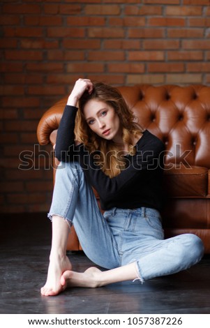 Young woman in jeans and pullover sitting on floor near brown leather sofa