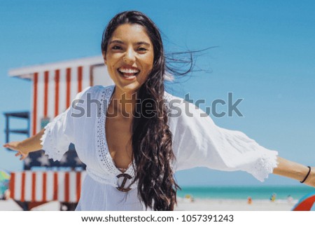 Young excited girl in white dress laughing happily at camera having fun on sunny tropical beach on background of blue sky.