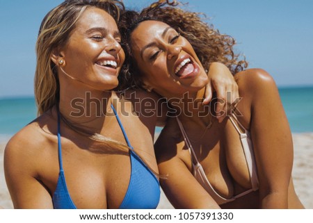 Laughing multiracial women in bikini embracing and having fun while chilling on sandy beach of ocean in bright sunshine.