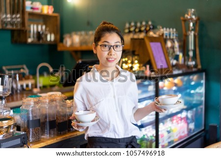 Cheerful pretty Asian waitress carrying coffee cups and looking at camera. Smiling attractive young coffee shop owner working in her cafe. Small business concept