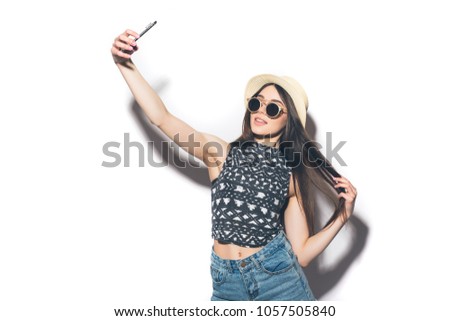 Young cheerful attractive brunette is smiling taking selfie on the camera of her phone, wearing casual summer outfit and a hat