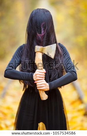 a girl in a witch costume in a long black dress with long black hair covering her face holds an ax in her hands. Day, autumn, a tunnel of yellow autumn leaves.thematic photossesion