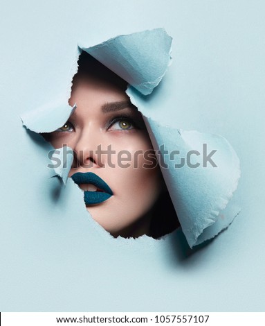 the face of a young beautiful girl with a bright make-up and puffy blue lips peers into a hole in blue paper.
