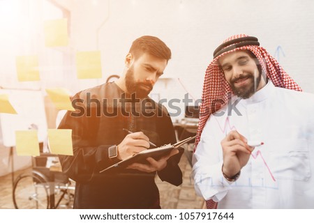 Two arab men in thawb working in office. Coworkers are taking notes in front of glass board.