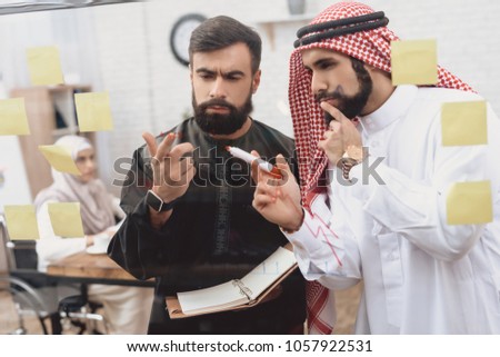 Two arab men in thawb working in office. Coworkers are taking notes on glass board.