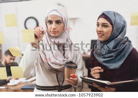 Two arab women in hijab working in office. Coworkers are taking notes on glass board.
