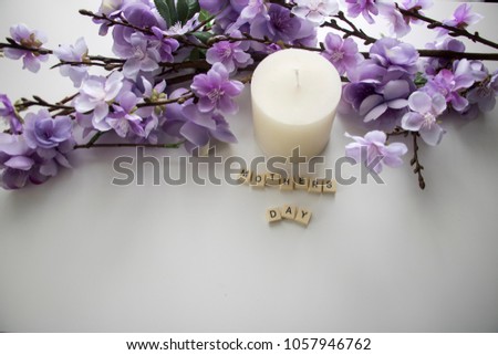 Block lettering placed together spelling mothers day on a white background and oval candle next to a bundle of purple flowers.