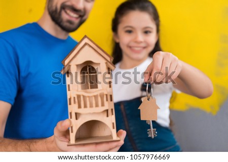 Bearded man holds toy wooden house in his hands and little girl holds keys to house. Concept of house selling. Moving to new house.