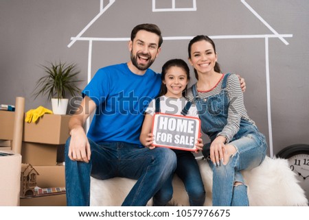 Real estate concept. Concept of house selling. Happy family sells house. Little girl is holding sign with inscription House for sale, sitting next to her parents.