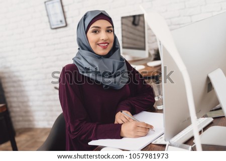 Arab woman in hijab working in office. Female worker is taking notes at table.