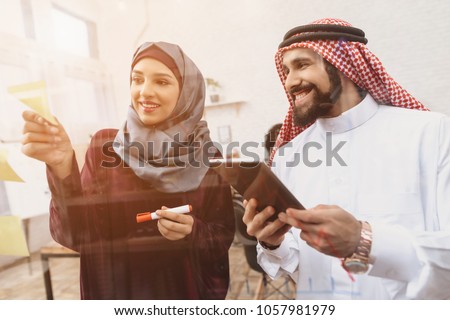 Arab man in thawb and woman in hijab working in offce. Coworkers are writing notes on glass board. Royalty-Free Stock Photo #1057981979