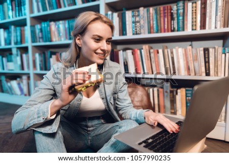 White girl sitting near bookshelf in library at night. Student is eating sandwitch and using laptop.