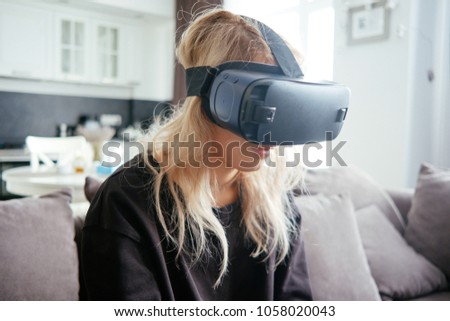 Young blonde girl in black shirt at home with modern design and kitchen put on virtual reality glasses and watch a movie or pictures