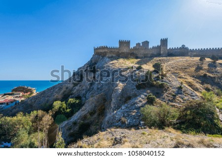 View on rocky slope with bushes and grass in close and ancient fortress with battlement wall and towers on mountain top in distance under clean blue sky in Crimea