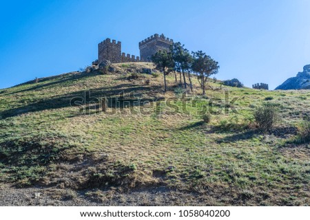 View on slope covered with green grass in close and few trees and ancient fortress with battlement wall and towers on mountain top in distance under clean blue sky in Crimea