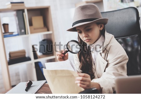 Private detective agency. Little girl in cloak and hat is sitting at desk looking at photos with magnifying glass. Royalty-Free Stock Photo #1058124272
