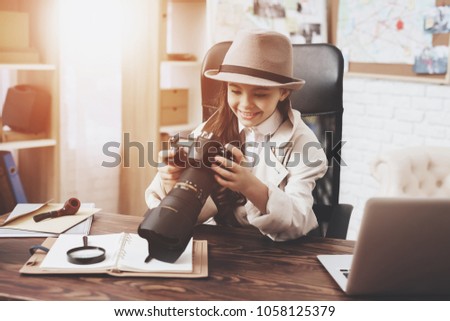 Private detective agency. Little girl in cloak and hat is sitting at desk looking at photos in camera.