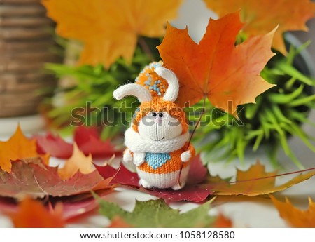 Handmade knitted toy. Easter Bunny in orange sweater and hat with pompom on the background of bright autumn maple leaves