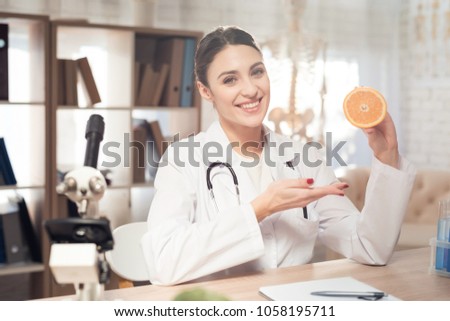 Female doctor in white gown sitting at desk in office with microscope and stethoscope. Woman is holding orange.