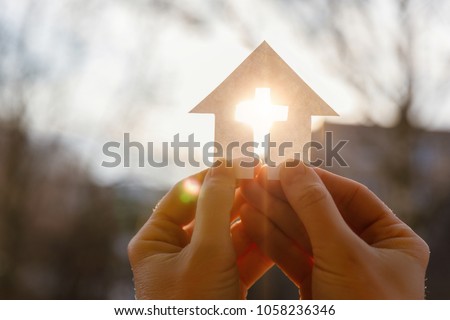 Hand with a house from paper and a cross on a background of sunrise.