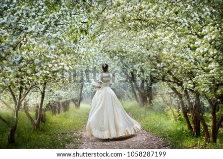 the girl in a white dress in the early morning in the Apple alley stands with her back and stretches her hand to the flowers