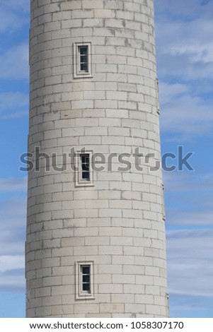 Close up outdoor view of part of a stone lighthouse. Round tower with vertical rectangular windows. Pattern of grey white stones blocks. Blue cloudy sky in background. Abstract architectural image.