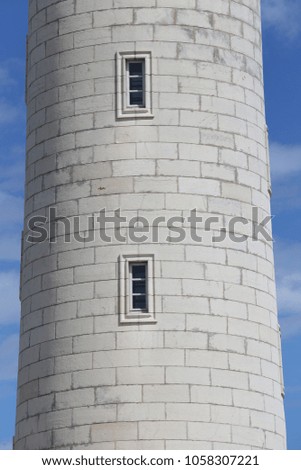 Close up outdoor view of part of a stone lighthouse. Round tower with vertical rectangular windows. Pattern of grey white stones blocks. Blue cloudy sky in background. Abstract architectural image.