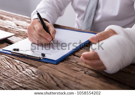 Close-up Of A Businessperson With Broken Arm Filling Health Insurance Claim Form On Wooden Desk Royalty-Free Stock Photo #1058554202
