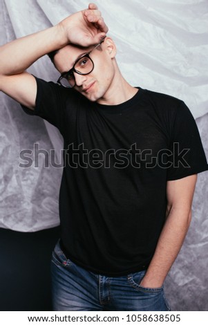 young guy brunette with short hair posing near a piece of fabric. on the clean face of a teenager glasses. emotional portrait of a student. streetwear style: spring t-shirt and skinny jeans