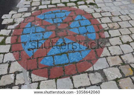 Close up outdoor view from above of a traffic sign painted on a old paved street and indicating: no parking. Pattern of square grey cobblestones. Circle and cross painted in red and with blue inside.