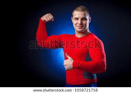 A strong dark-haired sportman  in a red sports wear  rush guard shows thumb up and biceps against a blue a lights on a black isolated  background 