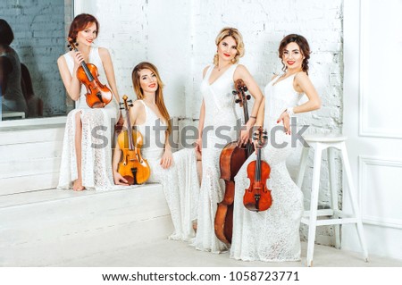 Beautiful girls posing with violins and a violoncello in white dresses in a bright interior room.