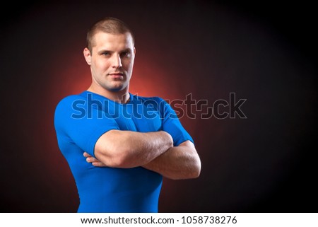 A strong dark-haired sportman  in a blue sports wear  rush guard  looks seriously at the camera and holds his arms crossed on his chest against a red a lights on a black isolated  background 