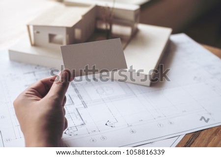A hand holding and showing an empty business card with an architecture model and shop drawing paper on table in office 