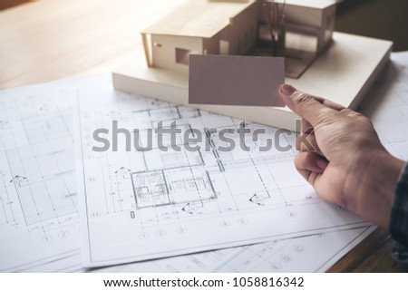 A hand holding and showing an empty business card with an architecture model and shop drawing paper on table in office 