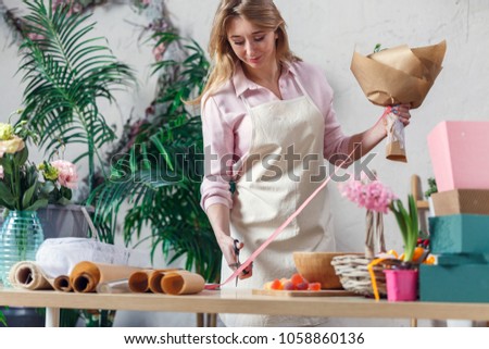 Picture of blonde florist in apron with bouquet at table with paper, marmalade, boxes