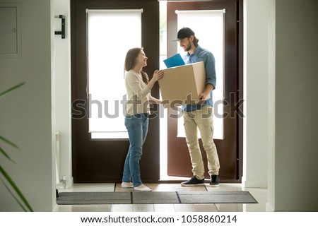 Happy young woman receiving parcel from smiling courier at home, delivery man holding carrying package giving cardboard box to customer receiver standing at doorway, door delivery service concept Royalty-Free Stock Photo #1058860214