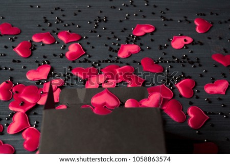 Red hearts and black rhinestones are poured out of a black gift box on a dark blue wooden table.