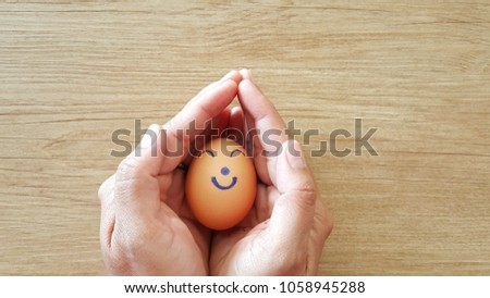 Two hands that support the egg on a wooden floor.
On the egg is drawn with a happy smile.
This picture is about caring for and loving someone you love.
Your loved ones are happy with your hands.