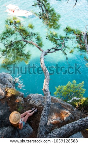 Girl in a straw hat sits on a rock under a tree. Woman looks at a boat in the sea. Top view of the pine branch on the rock. Tourist enjoys a beautiful view of the sea. Wild nature of Turkey, Kemer. Royalty-Free Stock Photo #1059128588